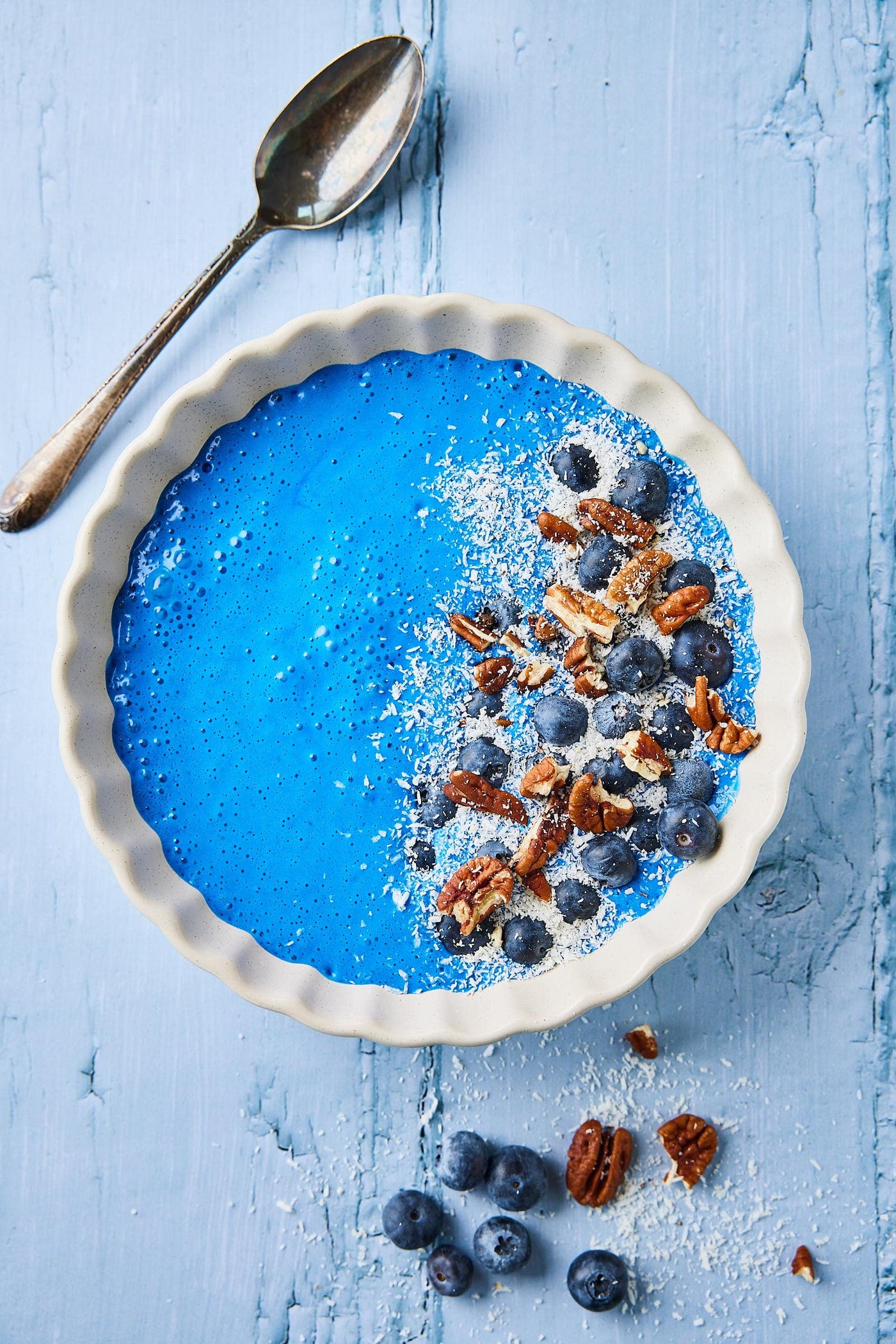 A bright blue spirulina smoothie bowl topped with blueberries, pecans, and shredded coconut, with a spoon on a pale blue wood background.