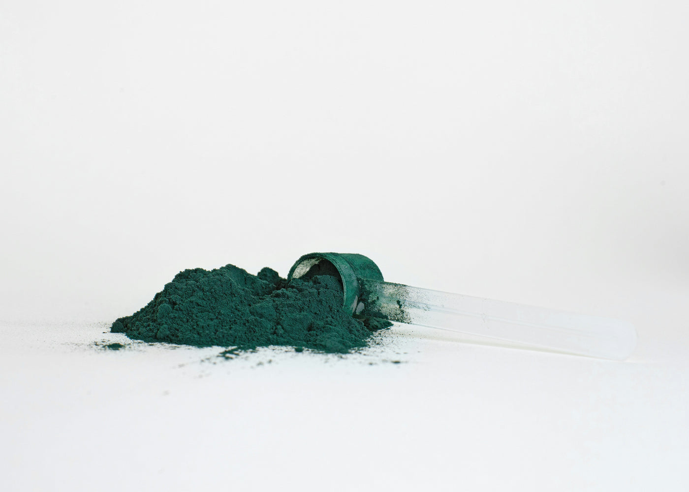 Deep green spirulina powder spilling from a measuring scoop beside a clear tube on a white background, roughly a single daily dose.